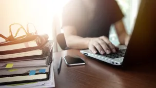 Man working on laptop at desk, with books and smartphone.