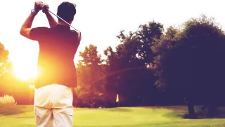 Golfer swings club, sunset in the background.