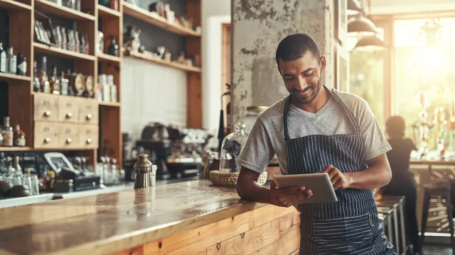Barista smiling, using a tablet, leaning on a bar.