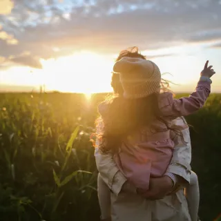 Mother carrying child on shoulders pointing to the sunset.